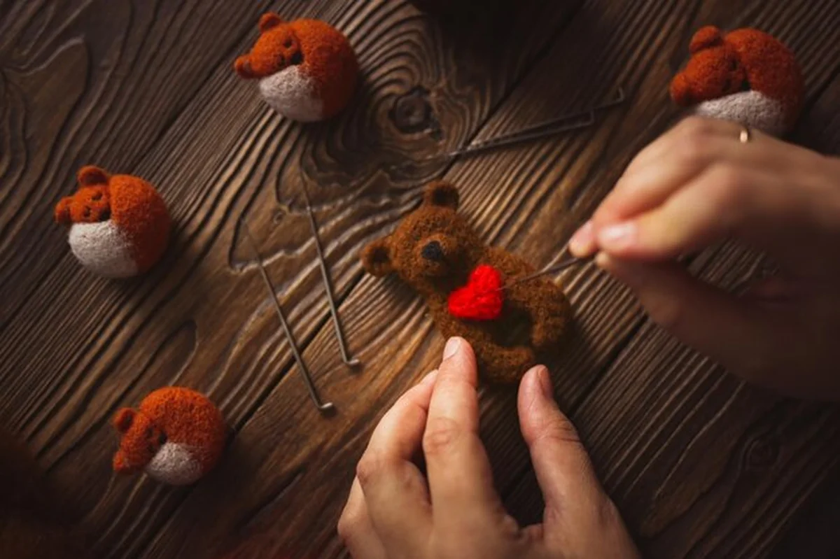 Ręce z igłami tworzą niedźwiedzia z sercem z wełny na drewnianym brązowym tle