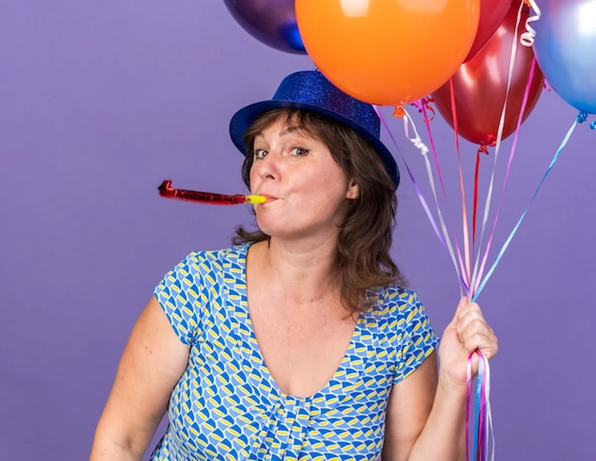 Kobieta w kapeluszu trzymająca balony dmuchane nad fioletową ścianą