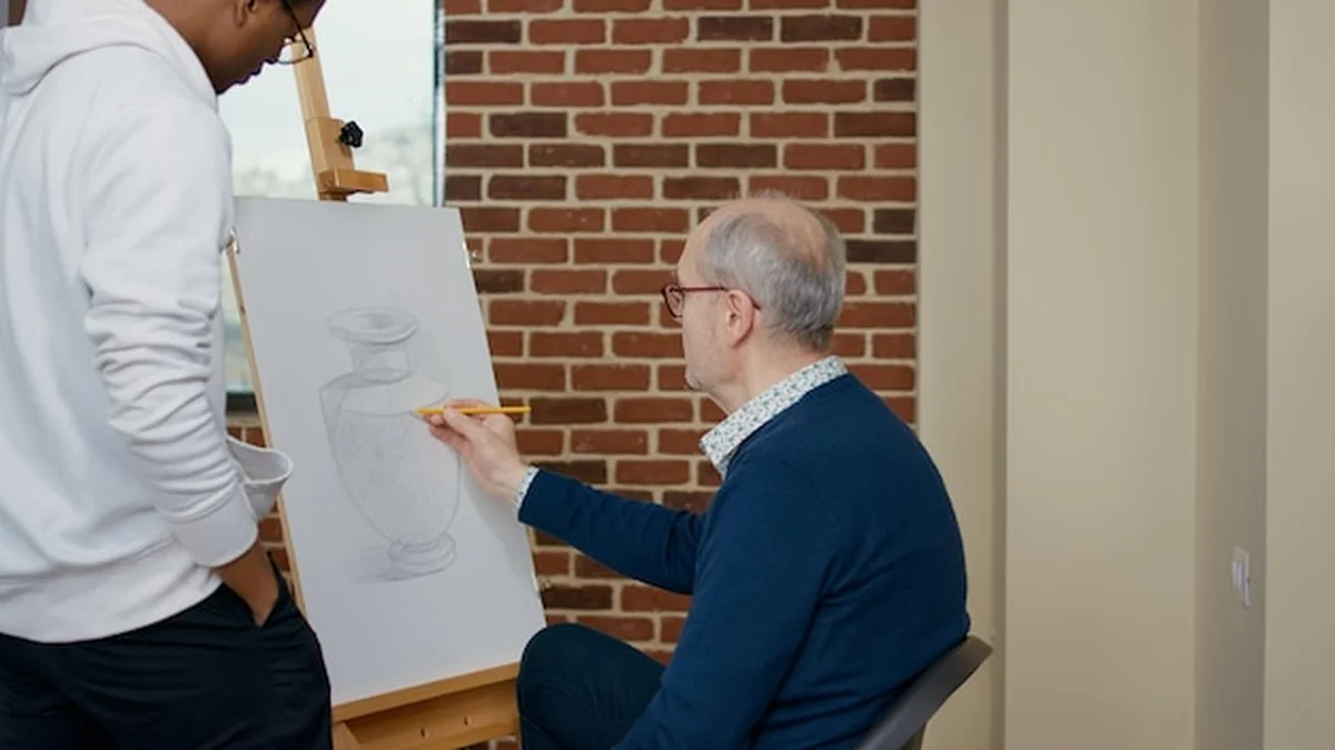 Osoba starsza rysująca z młodym nauczycielem na zajęciach plastycznych. Uczeń używa ołówka do szkicowania na płótnie